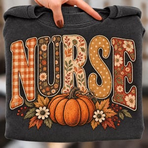 Può includere: Una felpa grigio scuro con la parola NURSE in grandi lettere stampatello. Ogni lettera presenta un motivo unico, tra cui quadretti vichy arancioni, stampe floreali, foglie autunnali e pois. Una zucca arancione si trova sotto con foglie.
