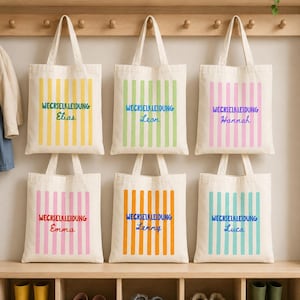 Op de afbeelding: Zes off-white canvas draagtassen hangend aan houten haken. Elke tas heeft verticale strepen in geel, lichtgroen, roze, rood, oranje of turquoise, met de tekst WECHSELKLEIDUNG en een unieke naam gedrukt in het midden van de strepen.