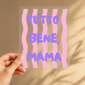 May include: A rectangular clear acrylic sign featuring vertical wavy light pink stripes and the text "TUTTO BENE, MAMA" printed in a bold, rounded purple font centered across the front surface of the transparent material.