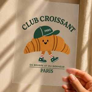 Könnte beinhalten: Ein durchscheinender rechteckiger Druck mit einem Cartoon-Croissant mit lächelndem Gesicht, das eine grüne Baseballkappe mit der Aufschrift PARIS und grüne Turnschuhe mit rot-weiß gestreiften Socken trägt. Der Text lautet CLUB CROISSANT, DU BEURRE ET DU BONHEUR, PARIS.