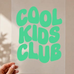May include: A rectangular, clear acrylic sheet featuring the text COOL KIDS CLUB printed in a bold, bright mint green color. The lettering uses a rounded, wavy, retro-inspired font style that fills the center of the transparent surface.