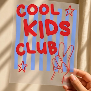 May include: A transparent rectangular sheet featuring the red text COOL KIDS CLUB, two red outlined stars, and a red line-art hand making a peace sign. The background of the sheet consists of vertical light blue stripes printed on the clear material.