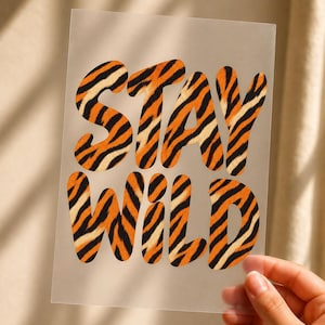 Könnte beinhalten: Ein rechteckiges, halbtransparentes Blatt mit dem Text STAY WILD in einer fetten, abgerundeten Schriftart. Die Buchstaben sind mit einem orange-schwarzen Tigerstreifenmuster gefüllt, was einen kontrastreichen Effekt auf dem Hintergrund erzeugt.