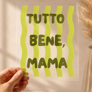 May include: A clear rectangular sheet featuring vertical wavy lime green stripes and the dark olive green text "TUTTO BENE, MAMA" printed in a bold, rounded sans-serif font. The design is centered on the transparent surface.