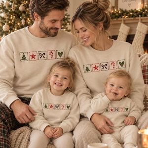 Puede incluir: Sudaderas beige a juego con cuello redondo que presentan una fila horizontal de cinco iconos cuadrados: un árbol de Navidad verde, una estrella roja, un lazo negro, un bastón de caramelo rojo y un corazón verde impresos en el pecho.