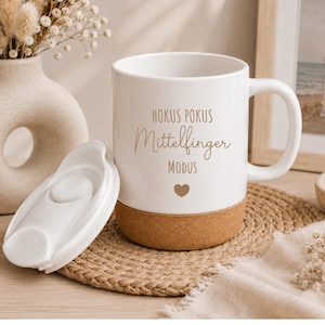 Könnte beinhalten: Weißer Keramikbecher mit Korkboden und weißem Deckel. Der Becher trägt die Worte "Hokus Pokus Mittelfinger Modus" in goldfarbenen Buchstaben sowie ein goldenes Herz. Der Becher steht auf einer gewebten Matte.