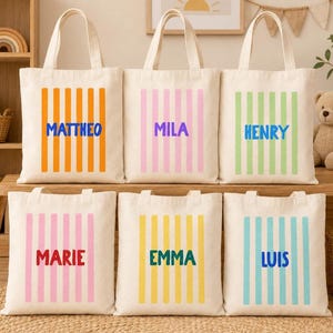 May include: Six off-white canvas tote bags arranged in two rows. Each bag features vertical stripes in orange, pink, light green, light pink, yellow, or light blue, with the names MATTHEO, MILA, HENRY, MARIE, EMMA, or LUIS printed in bold.