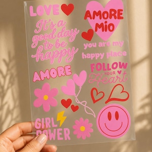 Könnte beinhalten: Ein transparenter Aufkleberbogen mit rosa und roten Motiven wie Herzen, Blumen, einem Smiley und einem gelben Blitz. Der Text umfasst LOVE, It's a good day to be happy, AMORE Mio, you are my happy place, AMORE, FOLLOW YOUR HEART und GIRL POWER.