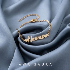 May include: A gold-toned metal name bracelet featuring the name Naomi in a cursive script font, flanked by two small heart shapes on either side. The bracelet is constructed with a delicate link chain and includes a lobster claw clasp closure.