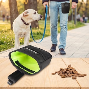 Könnte beinhalten: Schwarze Hundeleckerli-Tasche mit hellgrünem Innenraum, offen auf einer Holzoberfläche gezeigt. Ein Haufen Hundefutter liegt neben der Tasche. Eine Person geht mit einem gelben Labrador an einer blauen Leine im Hintergrund spazieren.