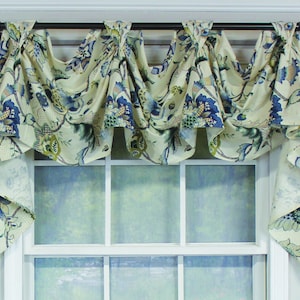 May include: Cream-colored window valance with a floral pattern in blue and green. The fabric is draped over a dark metal rod, creating a decorative, ruffled effect. The valance hangs above a white window frame.
