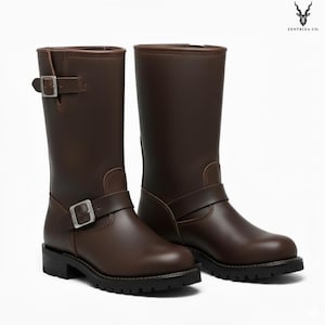 Puede incluir: Un par de botas de cuero marrones para motocicleta. Las botas tienen una altura media de pantorrilla, una puntera redondeada y un detalle de correa con hebilla. Las suelas son negras y las botas tienen un diseño robusto. Las botas se muestran sobre un fondo blanco.