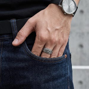 Op de afbeelding: Een zilverkleurige metalen ring met een herhalend reliëfpatroon van meerdere ogen rondom de band. De ring wordt gedragen aan een vinger, bij de zak van een donkerblauwe spijkerbroek, met een analoog polshorloge met zwarte band zichtbaar.