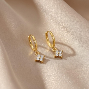 May include: A pair of gold-colored hoop earrings with a small, four-petal flower charm. Each charm is adorned with small, clear gemstones. The earrings are displayed on a satin-like, beige surface.