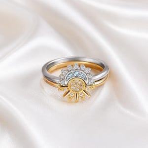 May include: A set of three rings with a celestial theme. The rings feature a gold band, a silver band, and a gold ring with a sunburst design. The sunburst is adorned with clear and light blue gemstones.