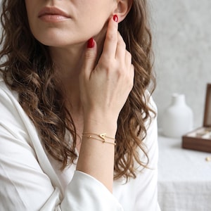 Op de afbeelding: Een verfijnde goudkleurige armband met dubbele ketting, voorzien van een bedel in de vorm van een hoofdletter A aan de bovenste ketting en een klein rond kraaltje aan de onderste ketting. De armband is gemaakt van dunne, gepolijste metalen schakels.
