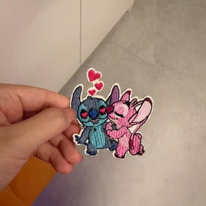 Puede incluir: Un parche bordado que muestra a un personaje azul con ojos en forma de corazón y a un personaje rosa con los ojos cerrados, ambos abrazándose. Sobre ellos hay tres pequeños corazones rosas. El parche tiene un borde blanco y está cosido con hilo vibrante.