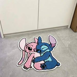 May include: A tufted rug featuring a blue character and a pink character embracing. The blue character has large ears and a dark nose, while the pink character has long ears and dark eyes. The rug has a white border and a textured, embroidered appearance.