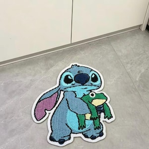 以下が含まれることがあります： 大きな耳と黒い鼻を持つ漫画のキャラクターが、黄色いお腹の小さな緑色のカエルを抱えている青い刺繍ワッペン。ワッペンには白い縁取りがあり、青と紫の色合いで細かくステッチが施されています。