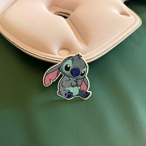 May include: An embroidered patch featuring a blue and grey alien character with large ears, a dark blue nose, and a light blue chest patch. The character is depicted in a seated position with its hands held together, outlined in a cream-colored border.