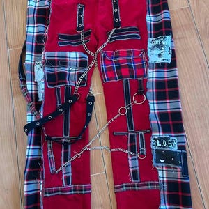 May include: Red punk-style pants with black straps, silver chain, and plaid patches. The pants feature multiple pockets and decorative metal rings. Patches with text and designs are sewn onto the pants.