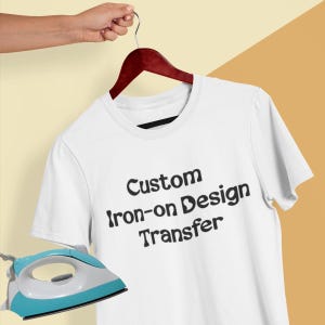 Può includere: Una t-shirt bianca a maniche corte appesa a una gruccia in legno scuro, con la scritta nera Custom iron-on Design Transfer stampata sul davanti. Un ferro da stiro azzurro e bianco è posizionato nell'angolo in basso a sinistra.