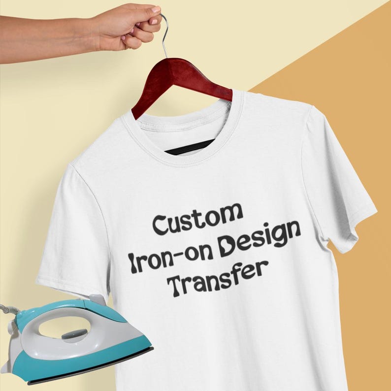 Może przedstawiać: Biały t-shirt z kr&oacute;tkim rękawem zawieszony na ciemnym drewnianym wieszaku, z czarnym napisem Custom iron-on Design Transfer nadrukowanym z przodu. Jasnoniebiesko-białe żelazko znajduje się w lewym dolnym rogu kadru.