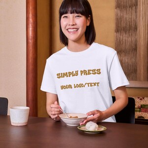 Może przedstawiać: Biały t-shirt z kr&oacute;tkim rękawem z brązowym napisem SIMPLY PRESS YOUR LOGO/TEXT nadrukowanym na klatce piersiowej. Koszulka posiada okrągły dekolt i standardowy kr&oacute;j, stanowiąc czyste tło dla własnych logo lub spersonalizowanych grafik.