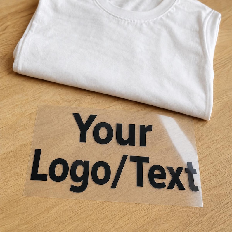 Może przedstawiać: Gładki biały t-shirt złożony starannie obok przezroczystego arkusza transferowego z czarnym napisem Your Logo/Text. Arkusz transferowy jest przeznaczony do nakładania na tkaninę, wyraźnie prezentując pogrubione litery na przezroczystej folii.