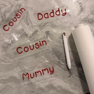 Może przedstawiać: Kilka przezroczystych naklejek winylowych z czerwonym napisem Cousin, Daddy, Mummy oraz Mary's Make up. Białe narzędzie do obierania z metalowym haczykiem oraz rolka białej taśmy transferowej znajdują się obok wyciętych liter z winylu.