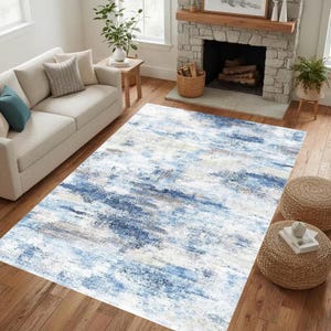 Könnte beinhalten: Ein rechteckiger Teppich mit einem abstrakten Design in Blau-, Weiß- und Grautönen. Der Teppich liegt auf einem Holzboden in einem Wohnzimmer. Der Raum umfasst ein weißes Sofa, einen Kamin und zwei geflochtene Hocker.