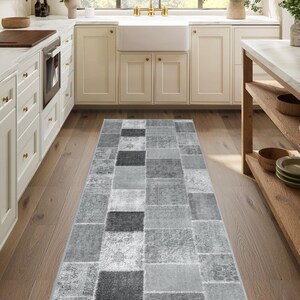Peut inclure: Un tapis rectangulaire de style patchwork dans des tons de gris et de blanc. Le tapis présente une variété de patchs carrés et rectangulaires avec différents motifs et textures. Le tapis est posé sur un sol en bois dans une cuisine.