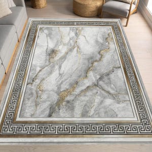 May include: A rectangular rug with a marble effect in grey and gold tones. It has a border with a Greek key pattern in black and gold. The rug is placed on a light-coloured wooden floor.