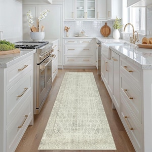 Può includere: Un tappeto runner rettangolare grigio chiaro e crema con un motivo vintage e usurato. Il tappeto è posizionato in una cucina con mobili bianchi, elettrodomestici in acciaio inossidabile e pavimenti in legno chiaro. Il design del tappeto presenta elementi geometrici e floreali.