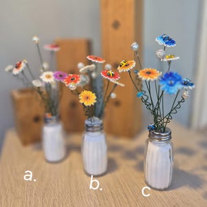 May include: Three decorative flower arrangements in repurposed salt shakers. Each arrangement features colorful, handcrafted flowers with wire stems and pearl accents. The flowers include daisies, sunflowers, and blue blooms, with some featuring small bee embellishments.