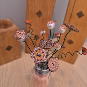 Op de afbeelding: Een decoratief arrangement van handgemaakte bloemen in een kleine glazen pot gevuld met roze korrels. De bloemen hebben verschillende knoopontwerpen met glitter- en kraalaccenten, tegen een houten achtergrond.