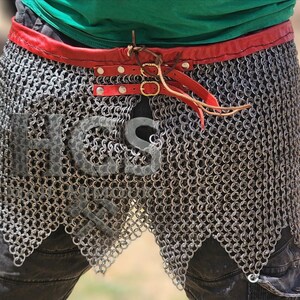 May include: A silver-colored chainmail skirt featuring a red leather waistband with two brass-toned buckles and leather straps. The chainmail is constructed from interlocking metal rings and has a scalloped hemline. The letters HCS are visible.