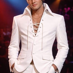 May include: A white suit with a lace-up front, wide lapels, and a high collar. The suit includes a matching jacket and pants. The person in the image is wearing a gold necklace. The background is blurred with red and blue lighting.