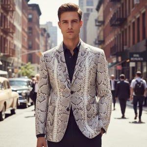 Puede incluir: Una blazer con estampado de piel de serpiente con cierre de dos botones, usado sobre una camisa negra. La chaqueta presenta una paleta de colores neutros de gris, blanco y negro. La imagen está tomada al aire libre en una calle de la ciudad.
