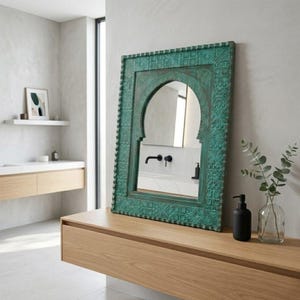 Op de afbeelding: Een turquoise, sierlijke spiegel met een gewelfde bovenkant staat op een licht houten oppervlak. De spiegel heeft gedetailleerde gravures. Een zwarte zeepdispenser en een heldere vaas met groen staan naast de spiegel. De spiegel reflecteert een badkamerinterieur.