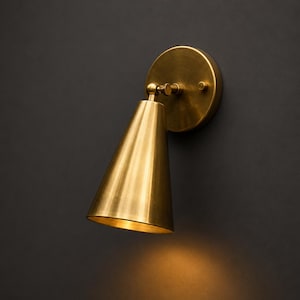 Op de afbeelding: Een wandlamp met een conische koperen kap en een ronde montageplaat. De lamp is voorzien van een verstelbaar draaigewricht dat de kap met de voet verbindt, afgewerkt in een geborstelde metallic goudtint met een warme lichtgloed.
