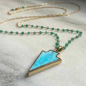 May include: A gold-colored necklace with a turquoise pendant in an arrow shape. The pendant is framed in gold and has a light blue stone. The necklace chain is gold with small green beads.