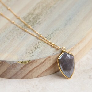 May include: A gold chain necklace with a dark gray faceted gemstone pendant. The pendant is shaped like a teardrop and has a gold setting.