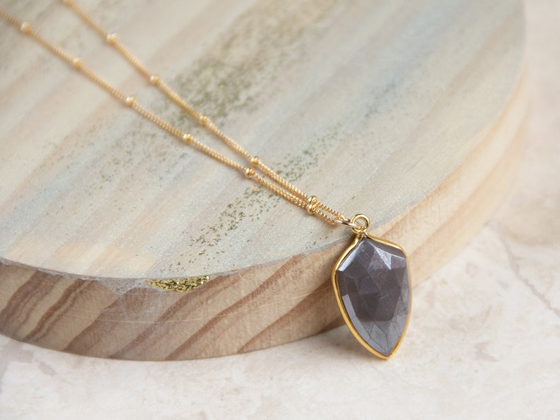 May include: A gold chain necklace with a dark gray faceted gemstone pendant. The pendant is shaped like a teardrop and has a gold setting.