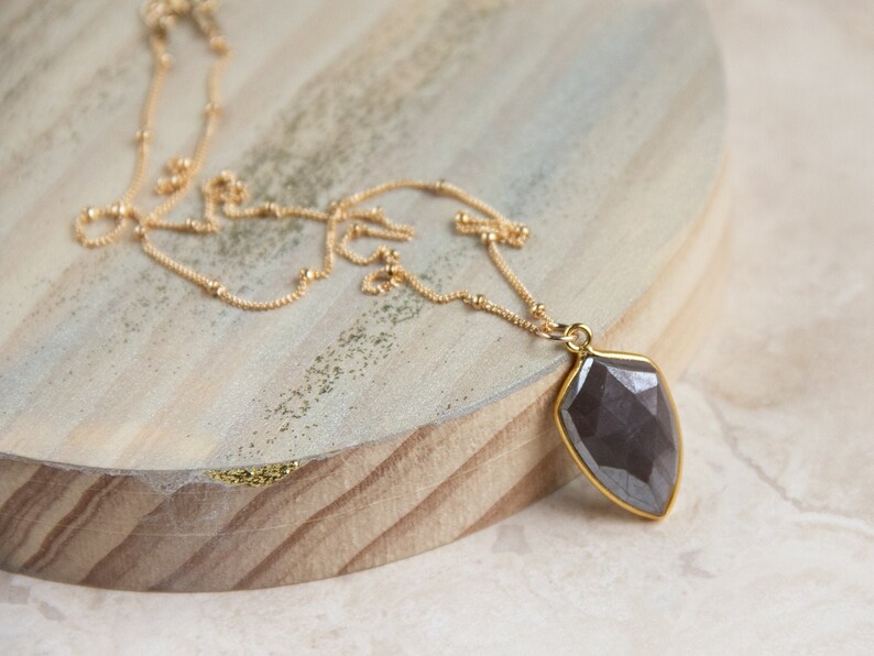May include: A gold chain necklace with a dark gray faceted gemstone pendant. The pendant is shaped like a teardrop and has a gold setting.