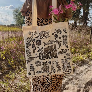 May include: A beige tote bag with a black print of various Florida wildlife and the words "SAVE FLORIDA." The bag is filled with pink flowers. The person is wearing a brown top and a leopard print skirt.