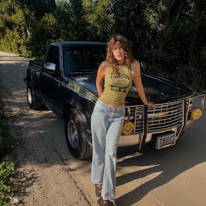 May include: A woman wearing a green tank top with the text "Florida Girl" and light blue jeans stands next to a black pickup truck. The truck has a license plate and two yellow smiley face lights on the front.