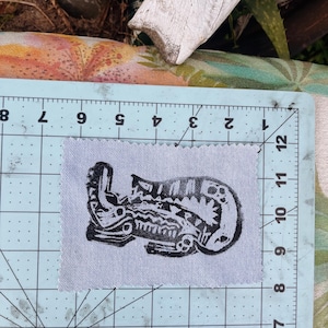 May include: A rectangular light gray fabric patch with pinked edges featuring a black ink block print of a stylized creature. The patch is positioned on a light blue cutting mat with visible inch markings, showing the patch is 4 inches wide.