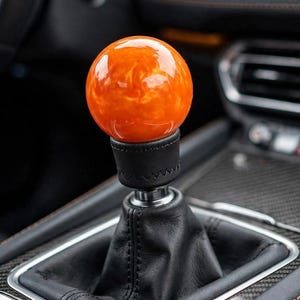 May include: A spherical gear shift knob with a glossy, marbled orange finish. The knob is mounted on a black leather boot with visible stitching, featuring a metallic silver collar connecting the sphere to the base of the gear shifter assembly.