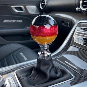 May include: A spherical gear shift knob featuring a horizontal tricolour design in black, red, and gold, mounted on a polished silver metal base with a black leather boot, installed in a vehicle interior with carbon fibre trim and AMG branding.